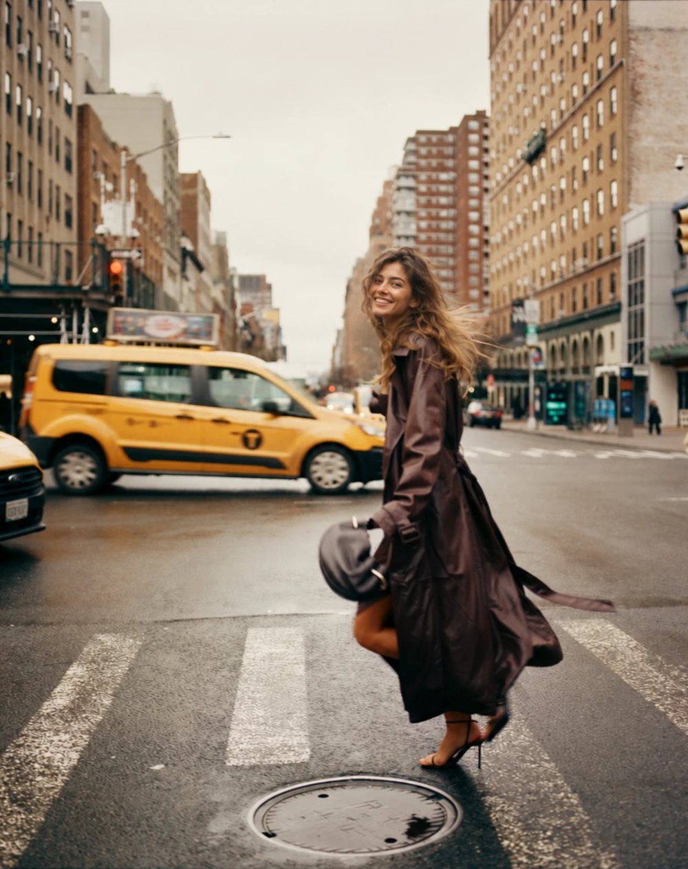 A woman in a long leather trench crossing a Manhattan street as a yellow cab passes